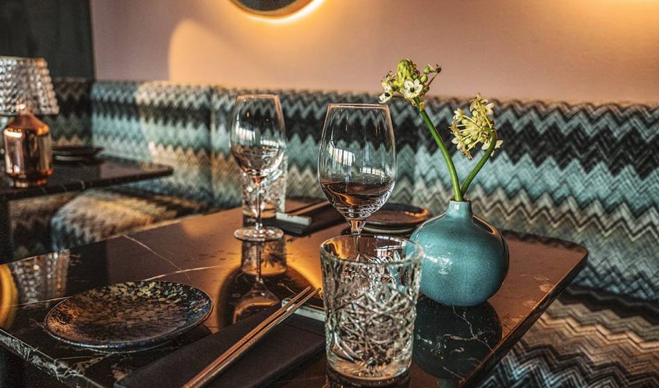 Detailed shot of a set table in #herrhoshi with crystal glass, wine glass and vase with flowers, in front of a patterned upholstered bench.