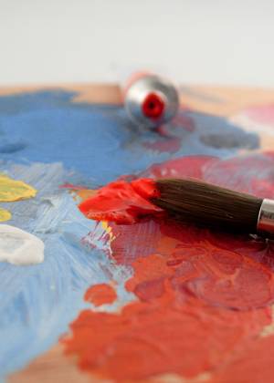 Eine Farbpalette mit bunten Farbflecken und ein Pinsel, der in rote Farbe getaucht wird, im Hintergrund eine Farbtube.