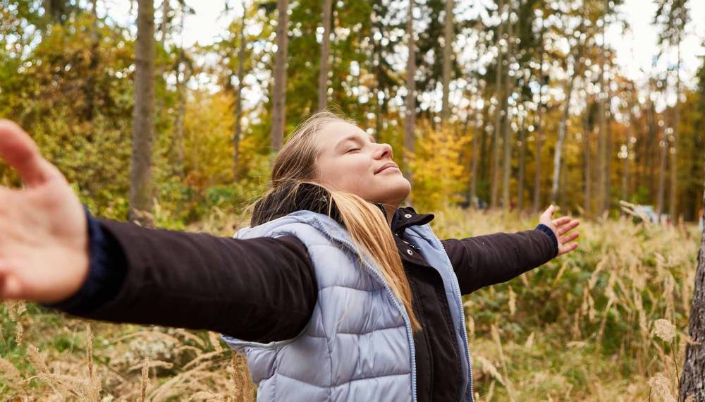 Eine Frau steht mit ausgestreckten Armen im Wald und macht bewusste Atemübungen inmitten der Natur.