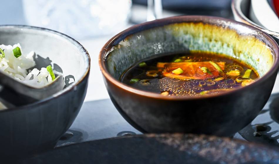 Close-up of a spicy soup with diced vegetables and herbs in a dark bowl, next to rice in #herrhoshi.