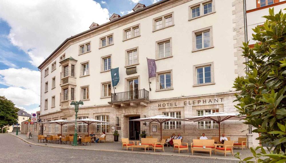 Außenansicht des Hotel Elephant Weimar mit Sitzmöglichkeiten, geöffneten Sonnenschirmen und Gästen.