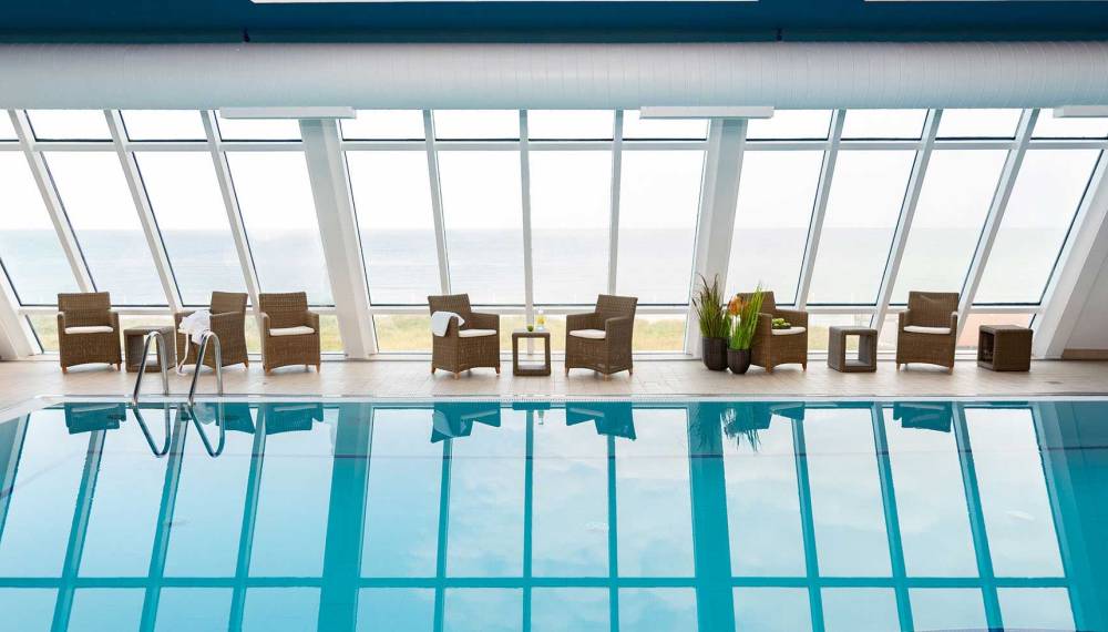 Heller Poolbereich im Wyn. Strandhotel Sylt mit Liegestühlen vor großen Fenstern, die einen Blick auf das Meer bieten.