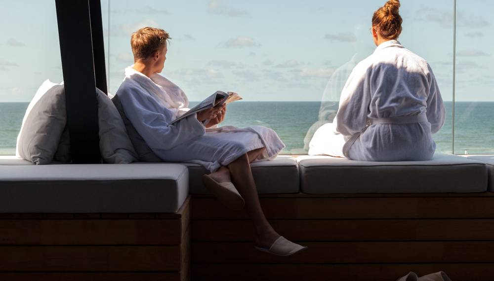 Ein Paar in weißen Bademänteln sitzt im Day Spa vor bodentiefen Fenstern mit Blick auf die Nordsee.