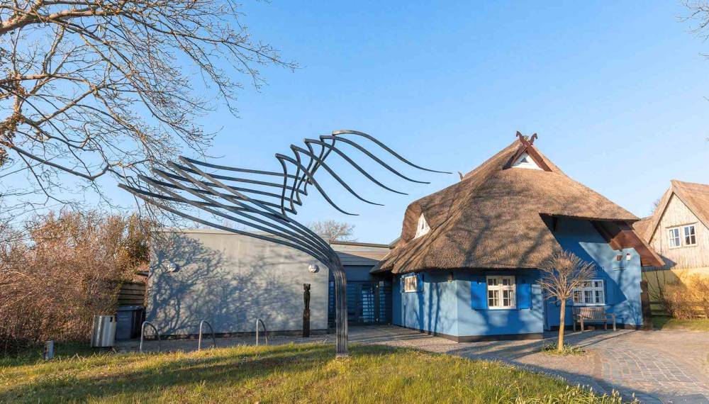 Außenansicht des Kunstkaten Ahrenshoop: Traditionelles Reetdachhaus in Blau, harmonisch kombiniert mit einem modernen Anbau.