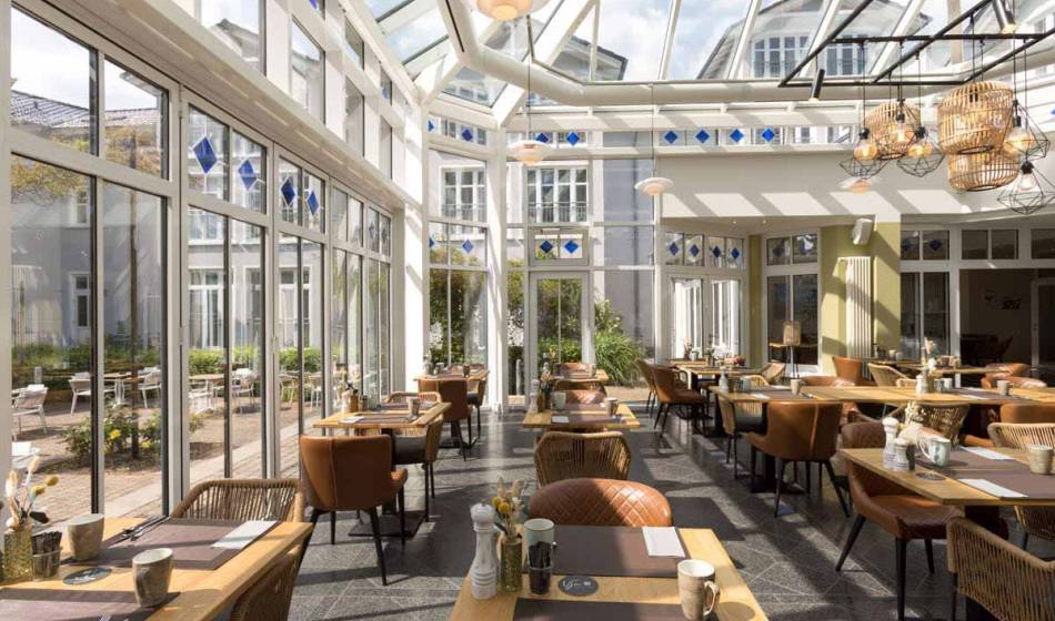 Light-flooded winter garden in the Vju Hotel Rügen, furnished with tables and chairs for breakfast.