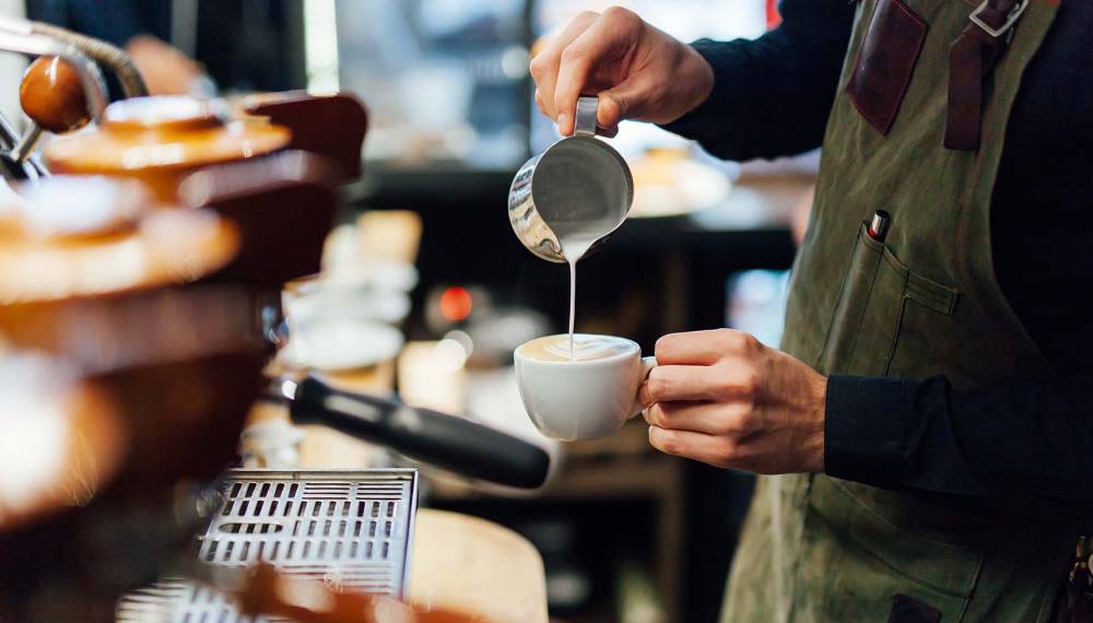 Ein Barista gießt geschäumte Milch in eine Tasse, um Latte Art zu kreieren, aufgenommen in einem gemütlichen Weimarer Kaffeehaus