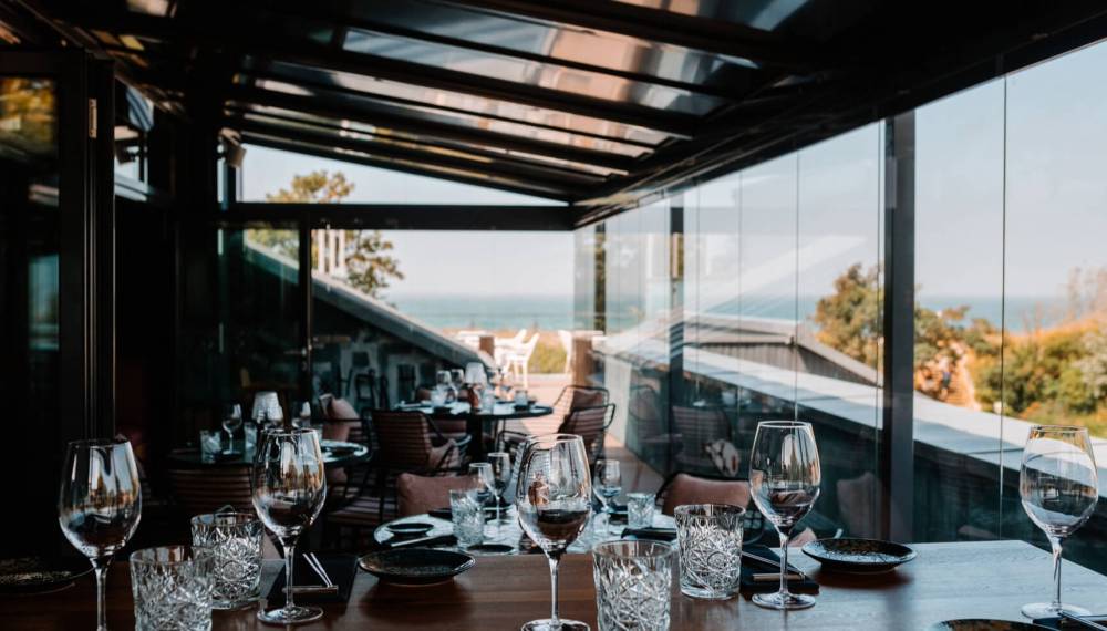 Restaurant #herrhoshi mit Glasfront und Meerblick, stilvolle Weingläser und Geschirr auf Holztischen in elegantem Ambiente.