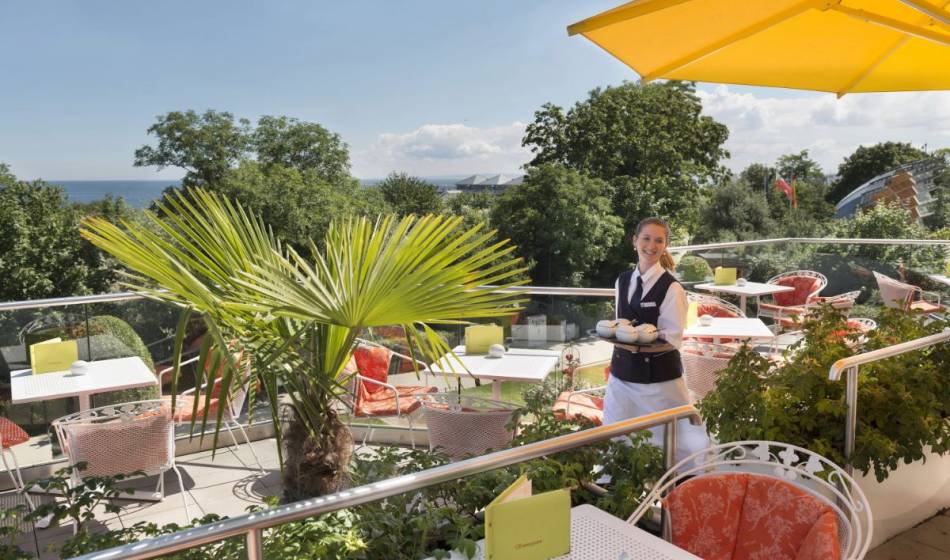 Sonnige Terrasse des Sommergartens im Kaiserhof Heringsdorf auf Usedom mit weißen Tischen, roten Stühlen, gelben Sonnenschirmen und tropischen Pflanzen. Eine Servicekraft trägt Getränke, während im Hintergrund Bäume und die Ostsee zu sehen sind.