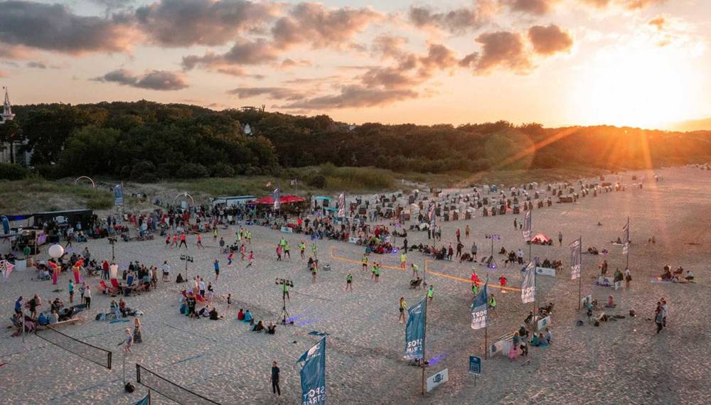 Belebter Ostseestrand bei Sonnenuntergang mit vielen Menschen, Beachvolleyballfeldern und Blick auf Dünenlandschaft.
