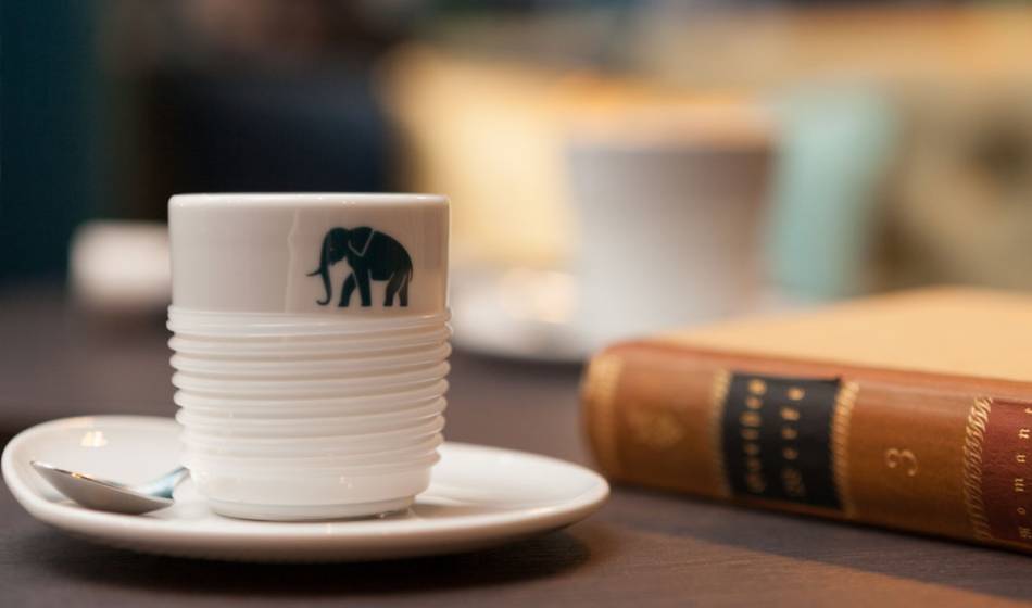 Weiße Kaffeetasse mit Elefanten-Logo auf Untertasse im Hotel Elephant Weimar, daneben ein Buch.