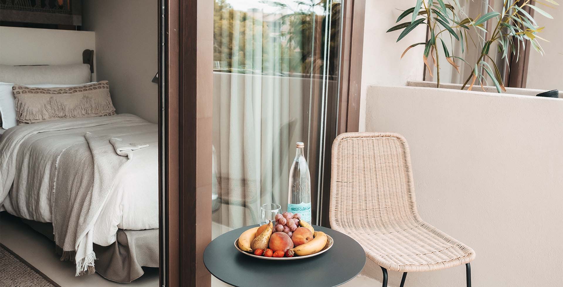 Zimmer Dormitorio im Barefoot Hotel Mallorca mit Bett, Balkon, einem Korbstuhl und Obstschale auf dem Tisch.