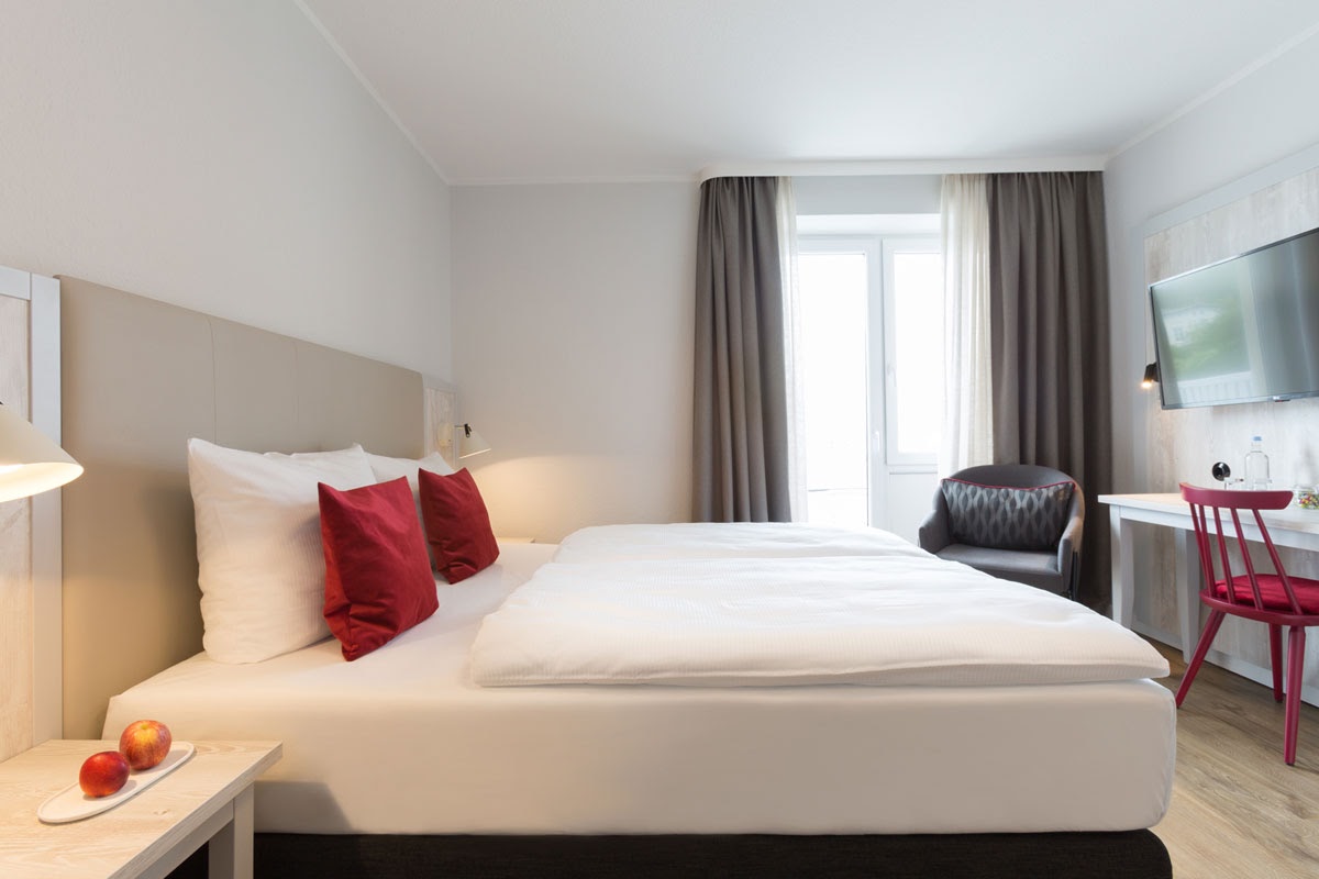 Helles Doppelzimmer im Koopmanns Hotel mit modernem Interieur, ausgestattet mit Bett, Sessel, Schreibtisch und großem Fenster.