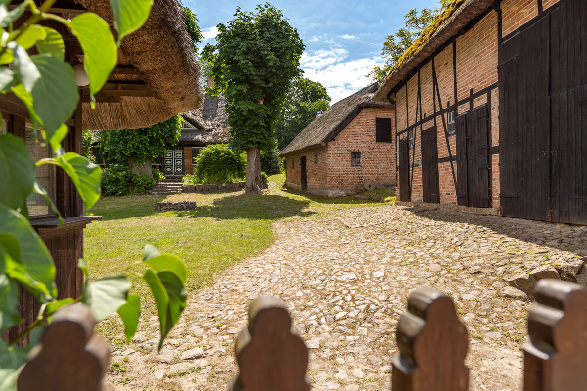 Historische Fachwerkhäuser mit Reetdächern, eingebettet in eine grüne Landschaft und sonnigem Innenhof mit Kopfsteinpflaster.