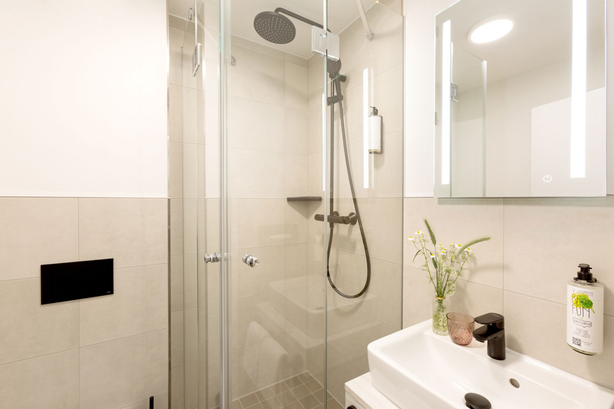 Badezimmer im Koopmanns Hotel &amp; Lädchen mit einer Regendusche, beleuchtetem Spiegel und Waschtisch.