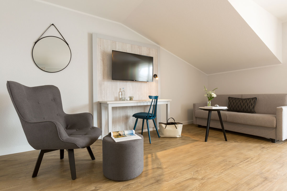 Sitzbereich eines Hotelzimmers im Koopmanns Hotel mit grauem Sofa, Sessel, Tisch, Schreibtisch und dekorativem Spiegel.