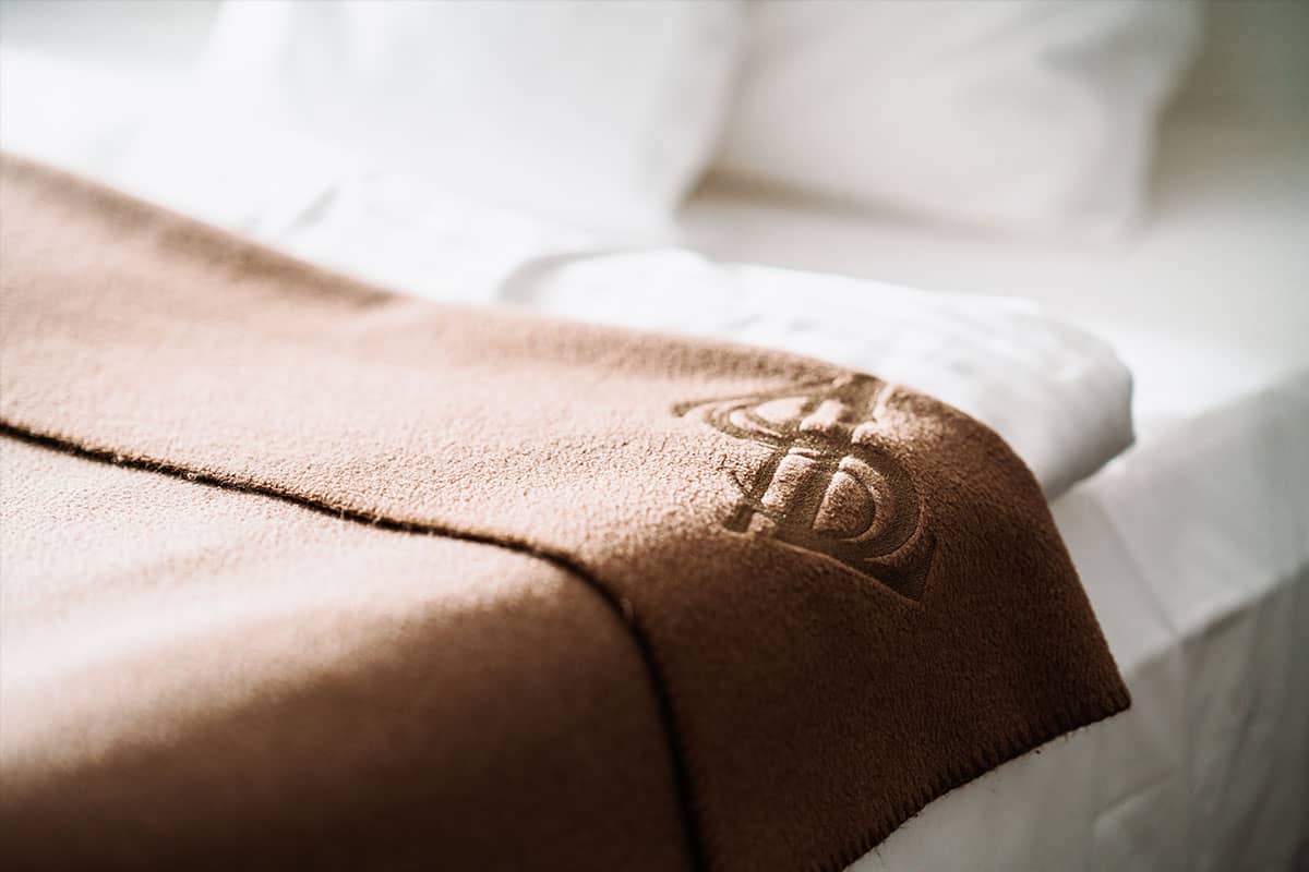 Brown blanket with hotel logo on a bed in the first-class double room of the Hotel Künstlerquartier Seezeichen.