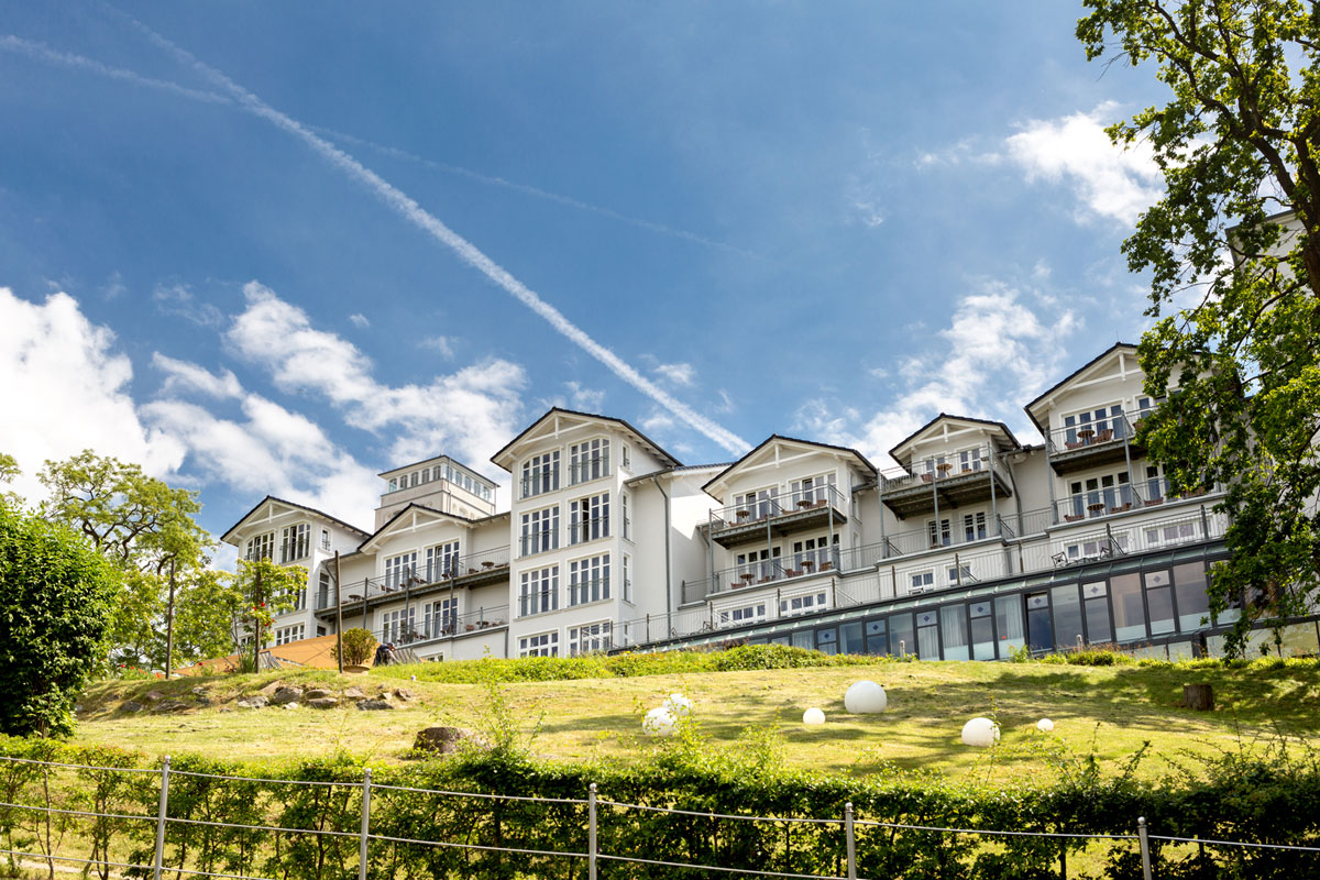 Außenansicht des Vju Hotels auf Rügen, eingebettet in grüne Natur, mit imposanter Architektur und blauen Himmel im Hintergrund.