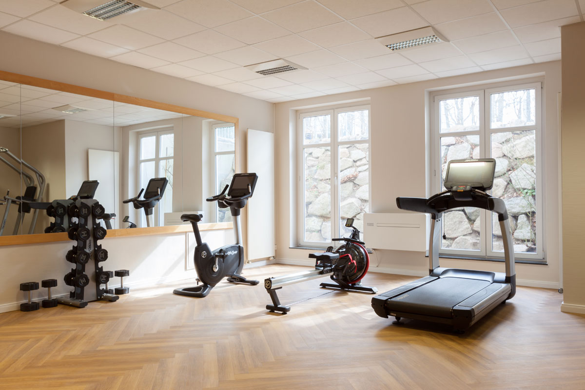 Moderner Fitnessraum im Vju Hotel auf Rügen mit Laufband, Fahrrad-Ergometer, Rudergerät und Hanteln vor großen Fenstern.