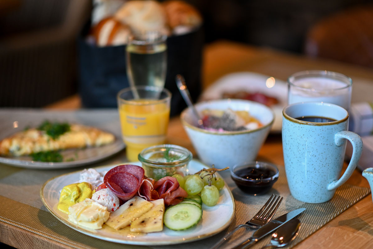 Frühstück im Vju Hotel Rügen mit frischem Obst, Käse, Wurst, Brot, Omelett, Joghurt, Kaffee und Orangensaft auf einem gedeckten Tisch.