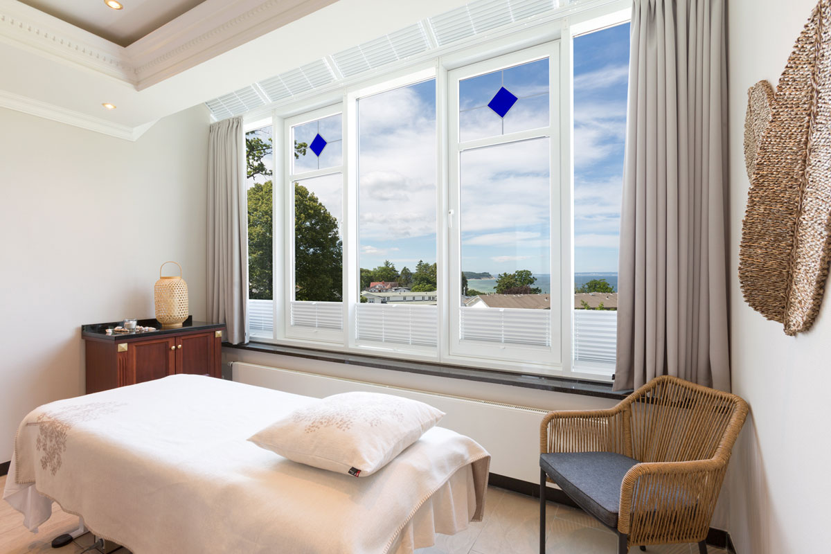 Behandlungsraum mit großen Fenstern, durch die man auf das Meer und die grüne Umgebung des Vju Hotel Rügen blickt.