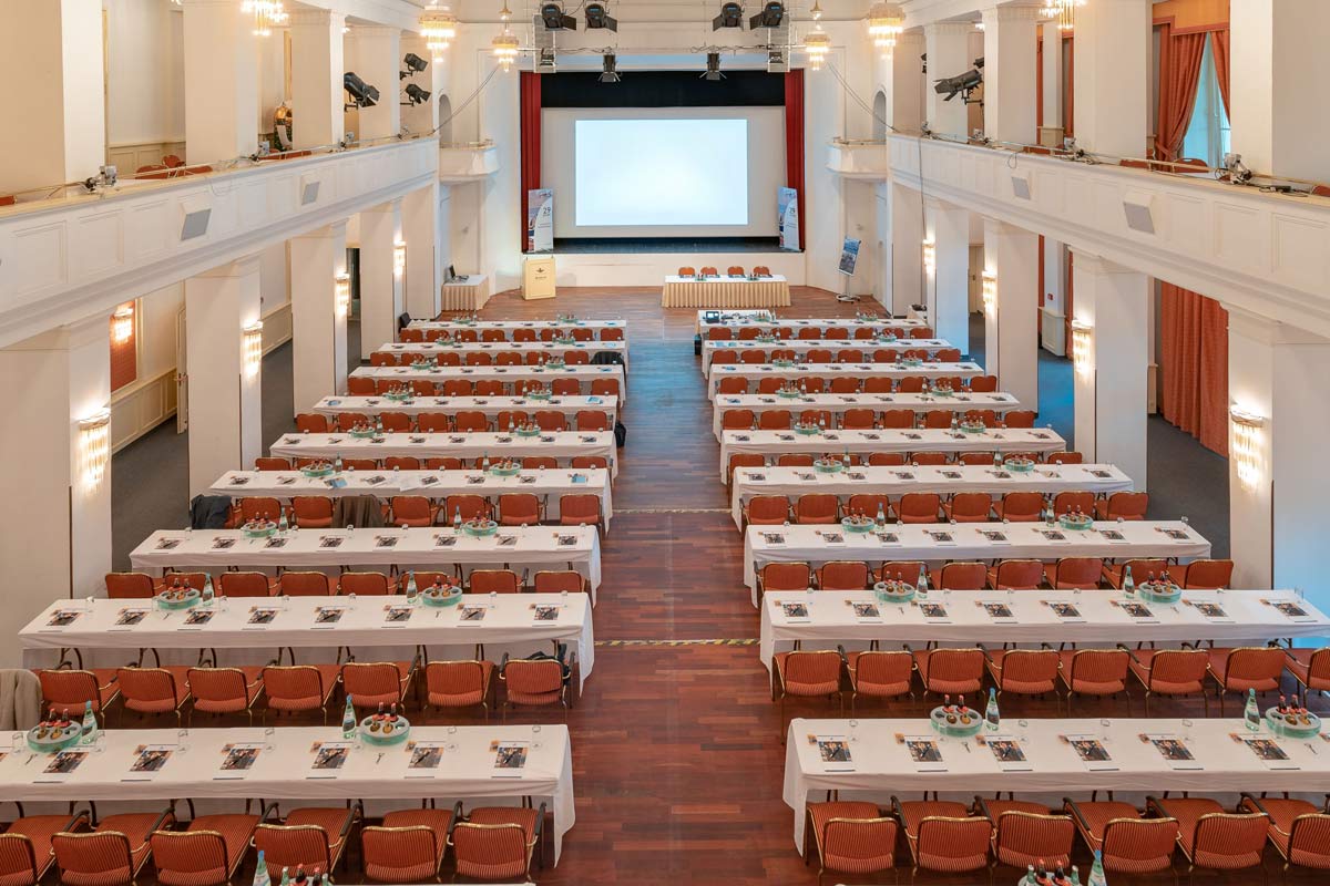 Kaiserbädersaal im Hotel Kaiserhof Heringsdorf, ausgestattet mit langen Tischen, Stühlen und einer großen Bühne.